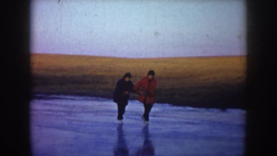 Two people skate hand in hand on a frozen lake in North Dakota, with a wide rural horizon behind them.