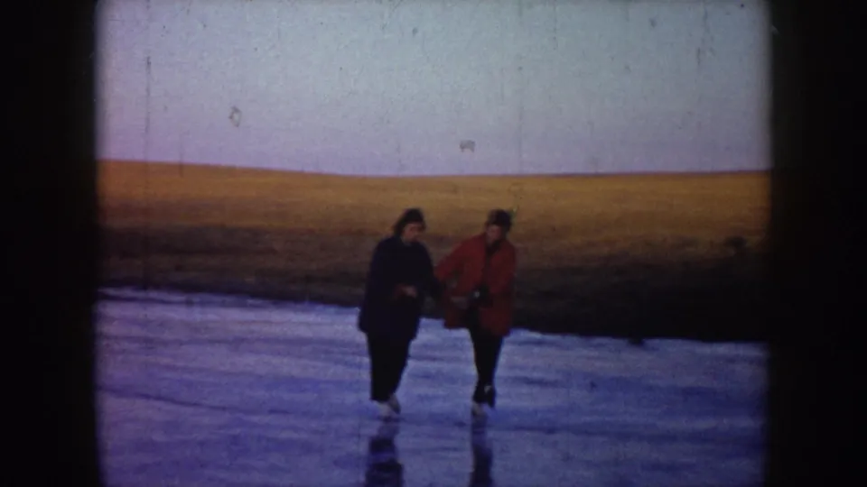Two skaters approach the camera on a frozen North Dakota lake, one wearing a red coat and the other a dark coat.
