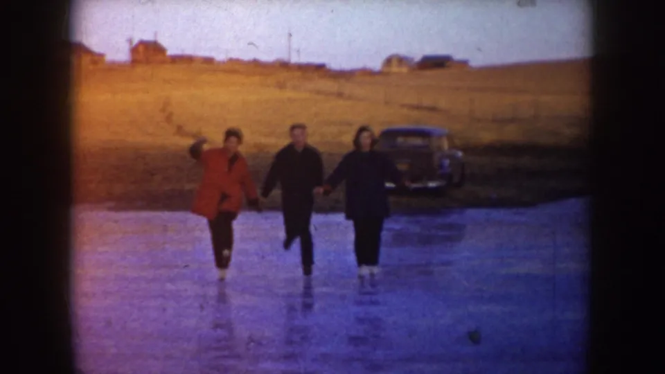 Three people skate hand in hand on a North Dakota frozen lake, with a dark parked car and distant buildings beyond the ice.
