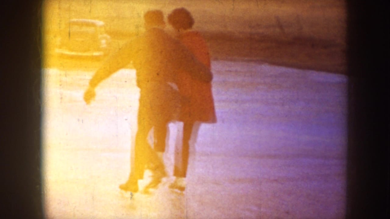 In supported 1953 North Dakota home-movie footage, two adults skate close together on a frozen pond near a parked car.