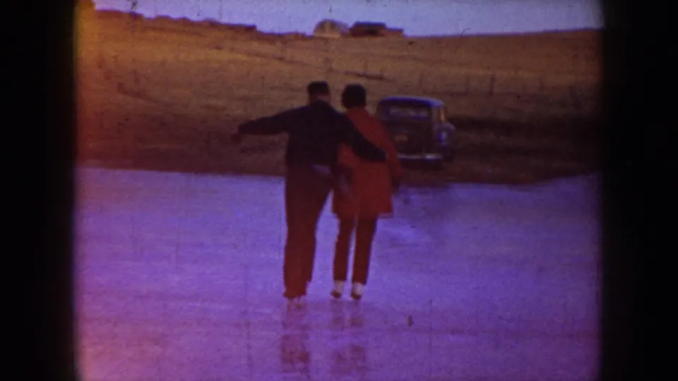 Two adults skate or pause together on a frozen pond in North Dakota, with an older parked car visible on the bank.