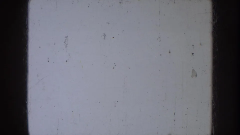 1953 North Dakota 8mm home-movie frame showing a pale blank film surface with dark gate edges, scratches, dust, and small debris.