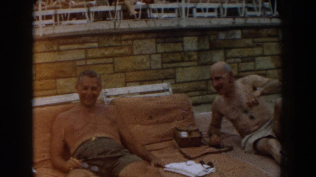 Two shirtless men relax on tan towels beside a stone wall and white lounge chairs.