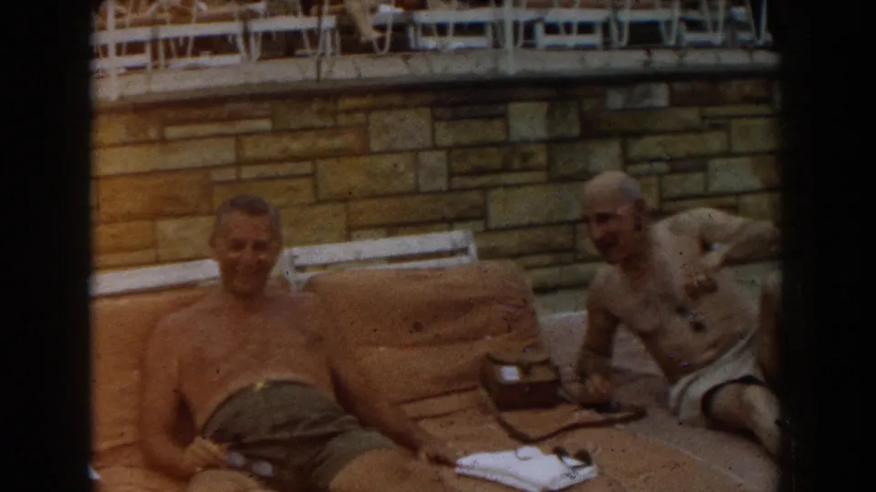 1961 Los Angeles home-movie frame showing two adult men lounging on tan poolside towels near a stone wall.