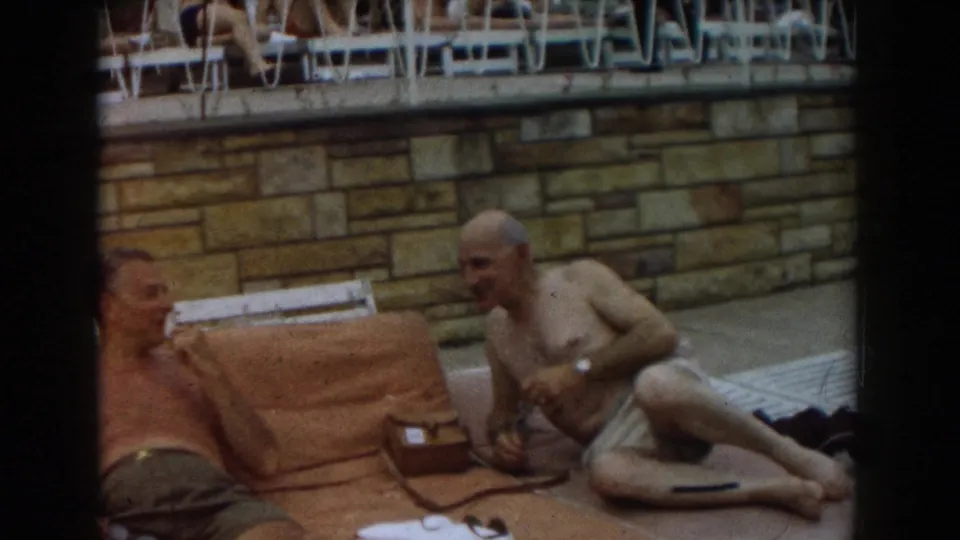 1961 Los Angeles poolside home-movie frame with one man reclining and another seated on a towel by a stone wall.