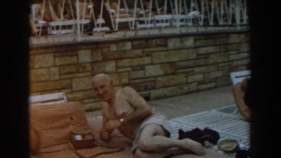 Poolside 1961 Los Angeles home-movie frame showing a man seated on a towel with clothing and a hat beside him.