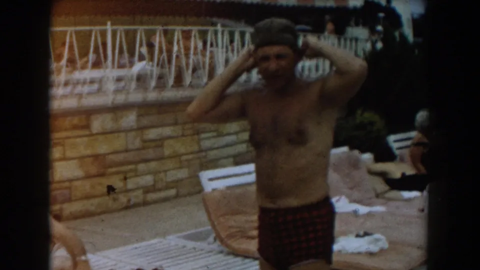 1961 Los Angeles home-movie frame showing a man in plaid swim trunks adjusting a swim cap near poolside towels.