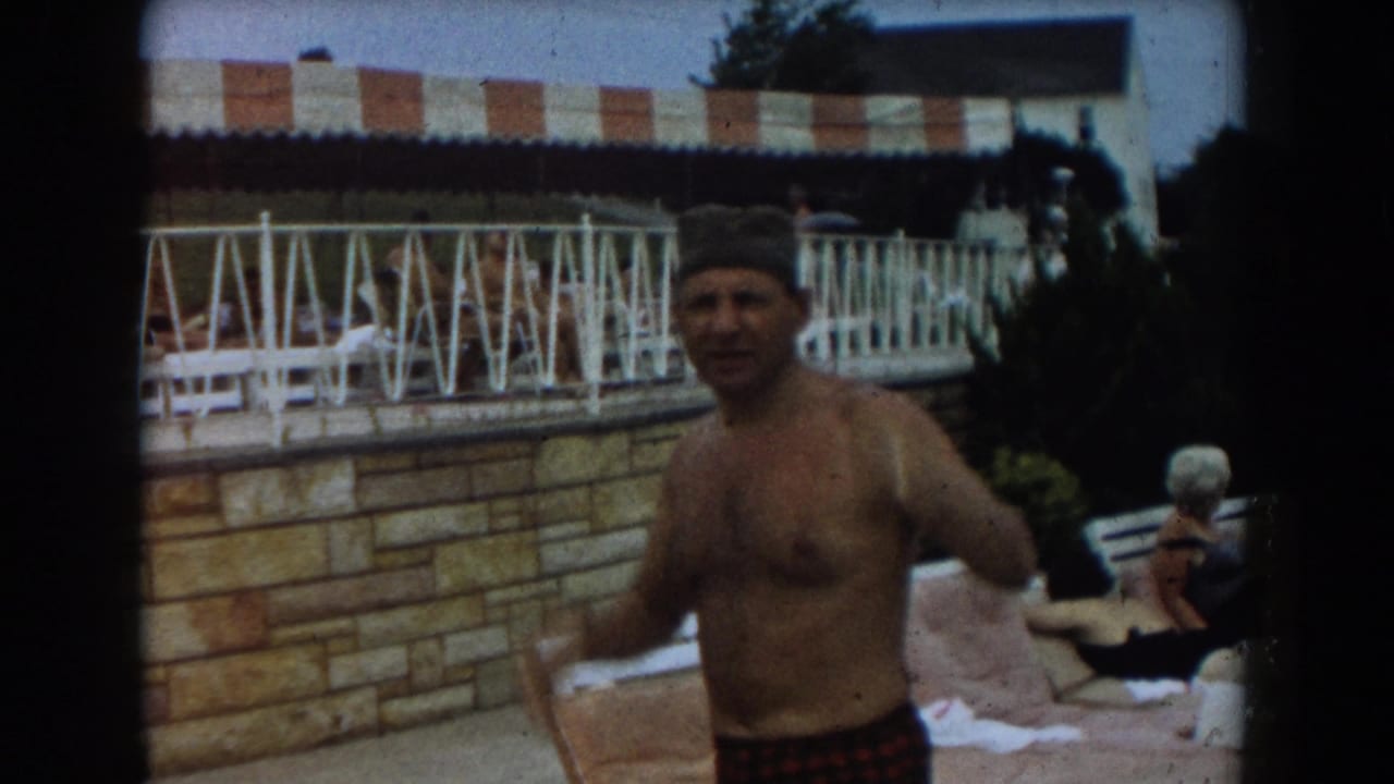 Man in swim cap and plaid trunks stands near an outdoor pool with lounge seating behind him.