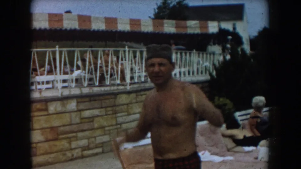In 1961 Los Angeles home-movie footage, a man in swim cap and plaid trunks stands poolside near white railings and a striped awning.
