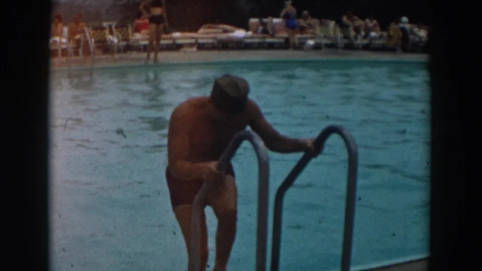 A man in a swim cap descends the pool ladder, framed by blue water, metal rails, and soft 8mm film grain.