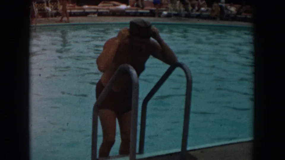 The swimmer pauses on the ladder and adjusts his dark swim cap, with the pool and lounge area behind him.