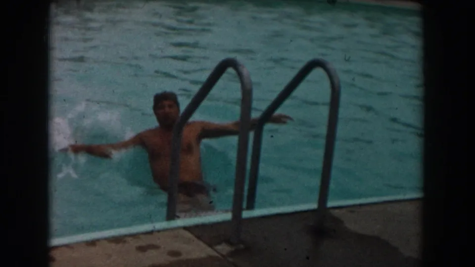A man in dark trunks splashes near the metal ladder, with the pool deck and blue-green water visible around him.