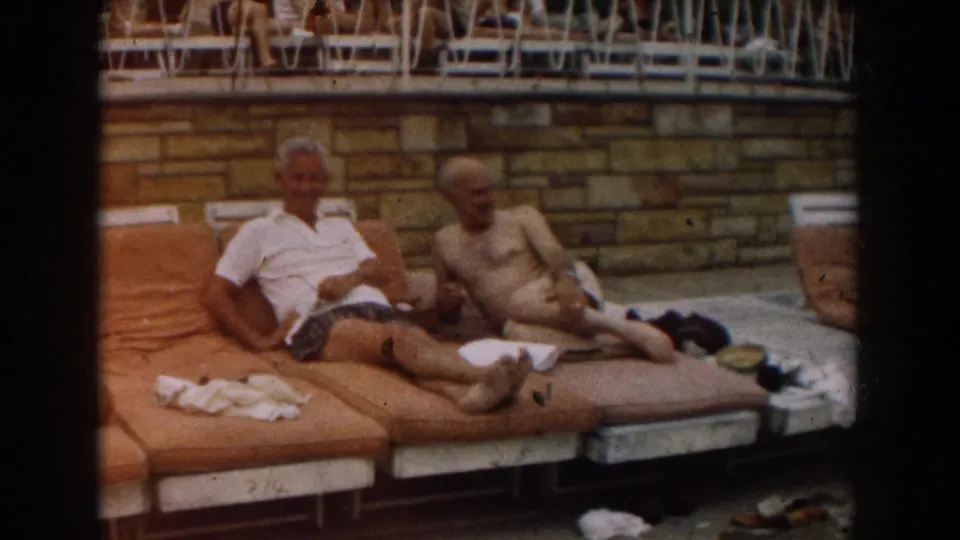 Poolside home-movie frame of two men on loungers in Los Angeles, 1961, with other guests visible on an upper deck.