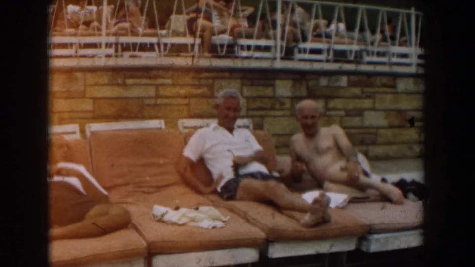 1961 Los Angeles poolside frame showing two older men reclining on orange-brown loungers near towels and a stone wall.
