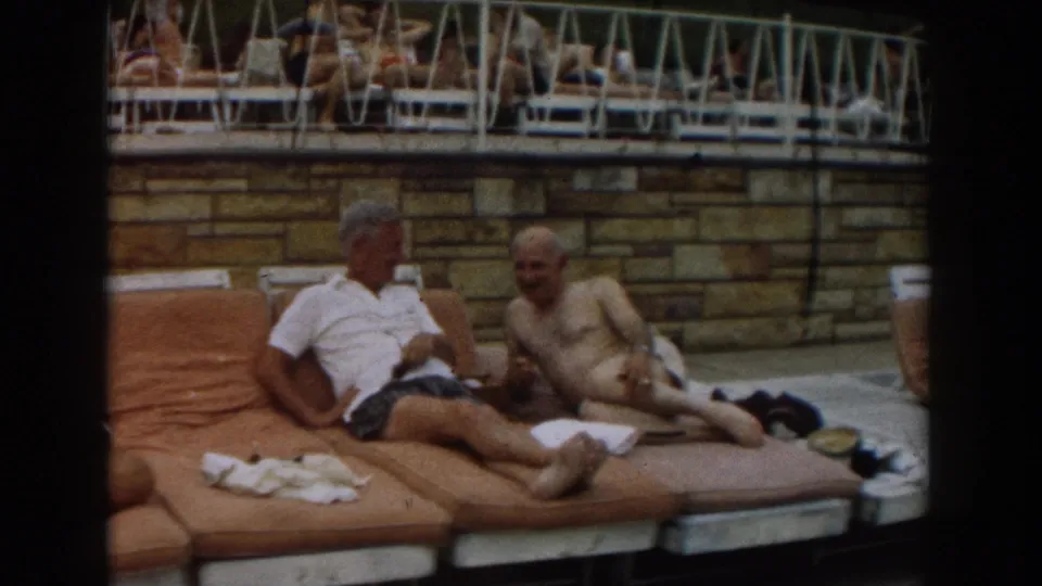 Late frame from a 1961 Los Angeles 8mm poolside scene with two men resting on adjacent cushioned loungers.