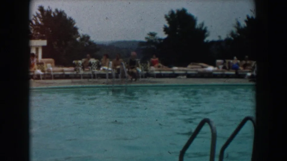 Outdoor pool view with seated guests, lounge chairs, trees, distant hills, and a pool ladder in 1961 Los Angeles home-movie footage.