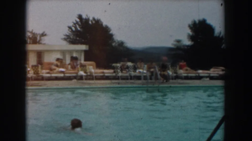 A swimmer moves through the foreground of a Los Angeles pool scene, with adults lounging and sitting across the deck in 1961 footage.
