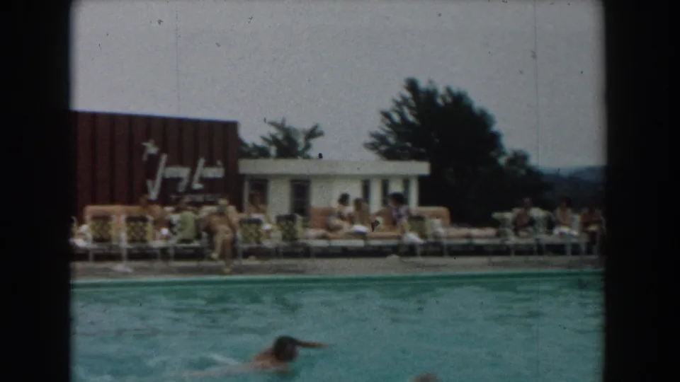 1961 Los Angeles poolside view with a swimmer, rows of lounge chairs, guests, trees, and a low building with a visible sign.