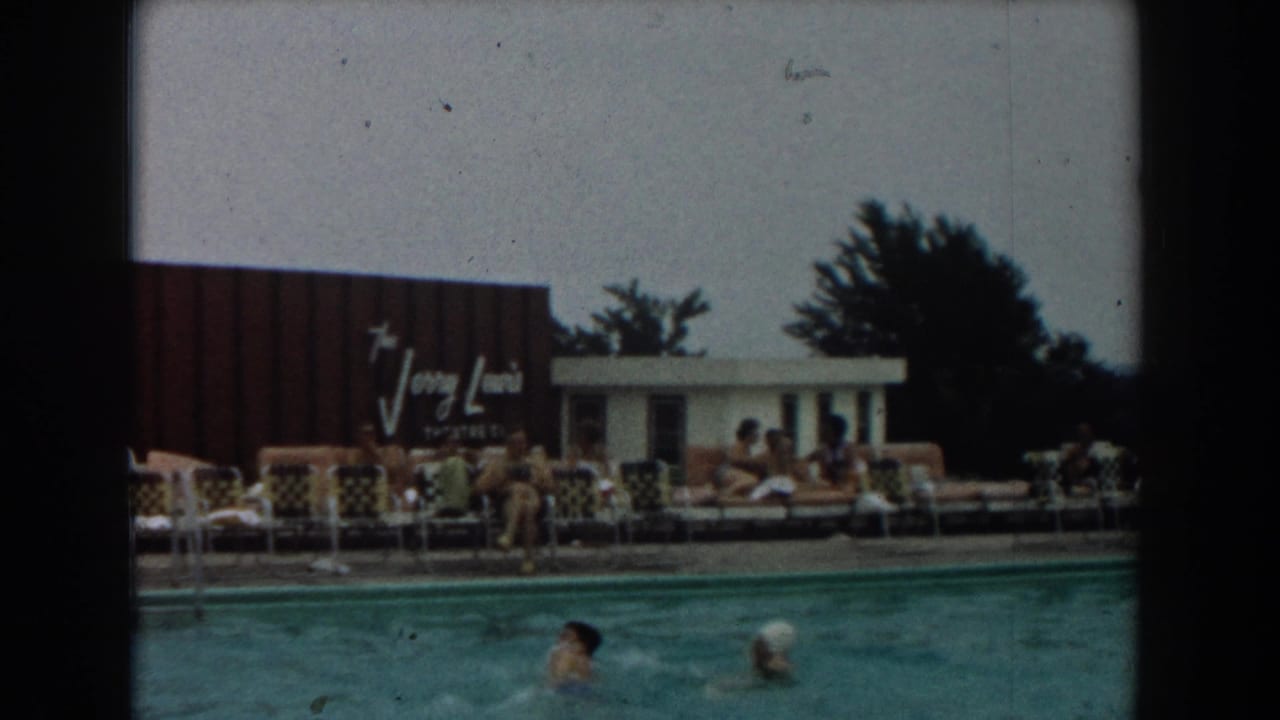 Swimmers in a pool with sunbathers on lounge chairs and a Jerry Lewis sign behind them.