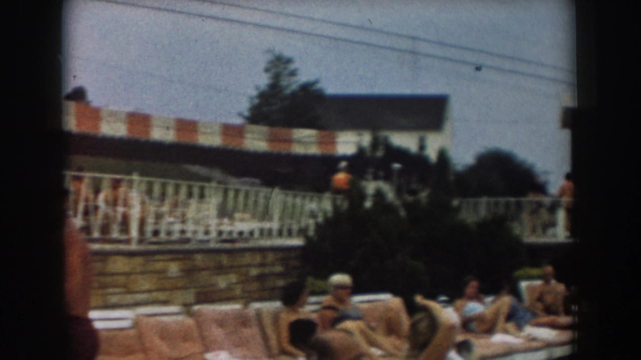 1961 Los Angeles poolside deck with adults seated behind a railing under a red-and-white striped awning.