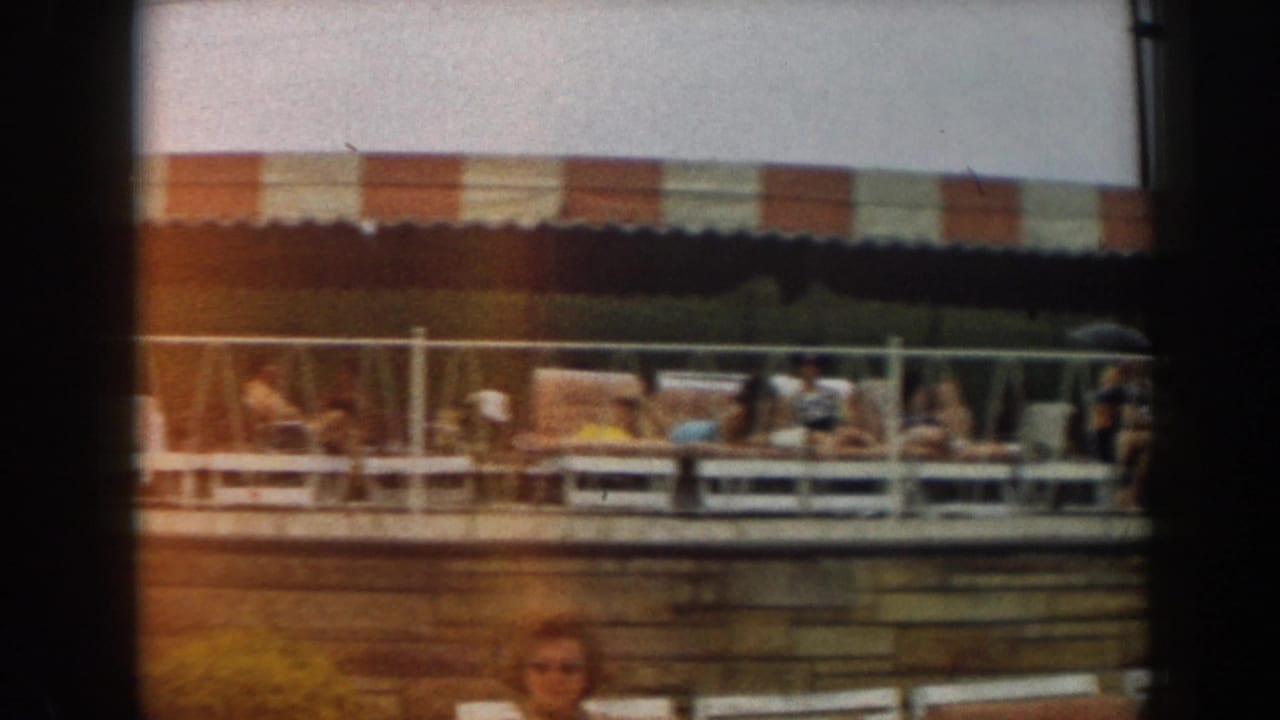 Poolside lounge area under a striped awning with adults seated behind a railing.