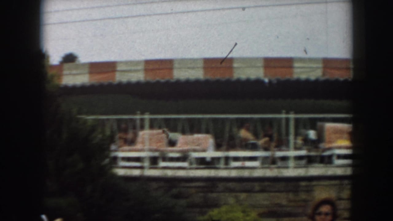 Lounge chairs and adults under a striped awning, partly obscured by dark film framing.