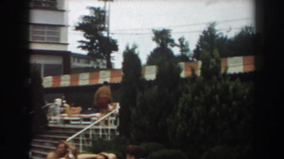1961 Los Angeles pool area with steps, evergreens, lounge furniture, and an adult moving near the shaded deck.