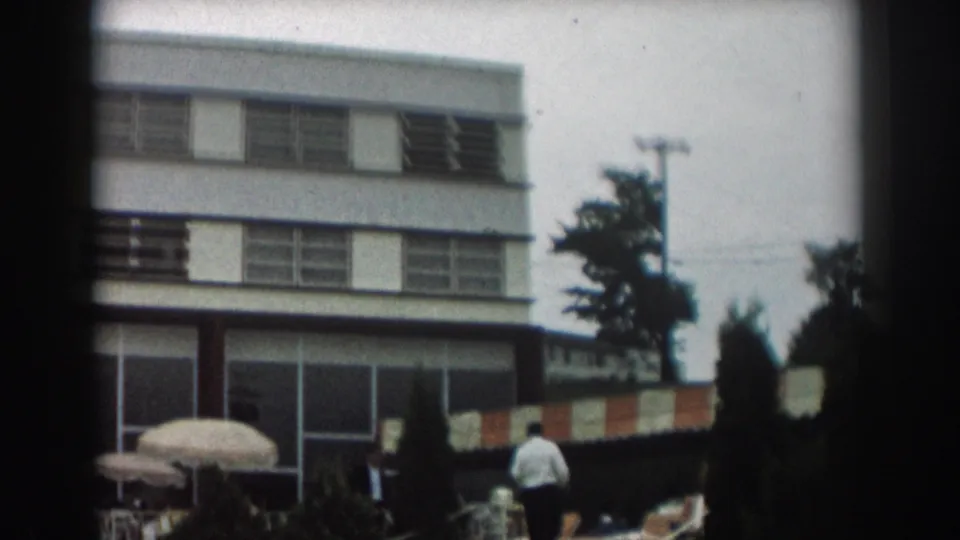 Los Angeles poolside view with a modern low-rise building, striped awning, patio umbrella, trees, and an adult walking nearby.