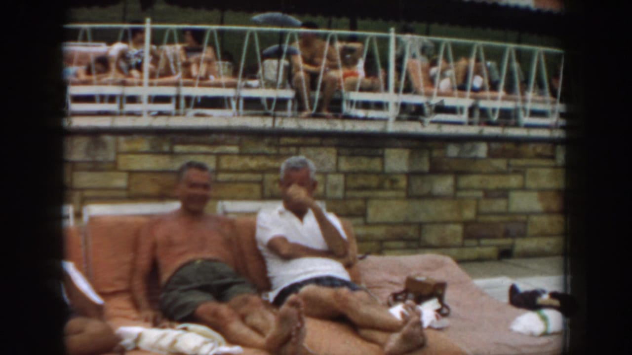 Two men lounge on orange poolside chairs below a raised deck with sunbathers.