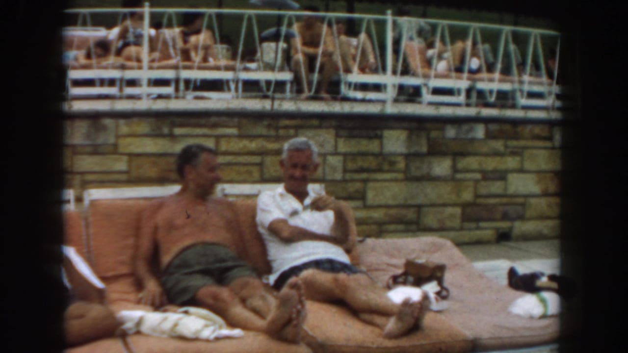 1961 Los Angeles frame of two men seated on orange poolside loungers, with a raised deck and other guests behind them.