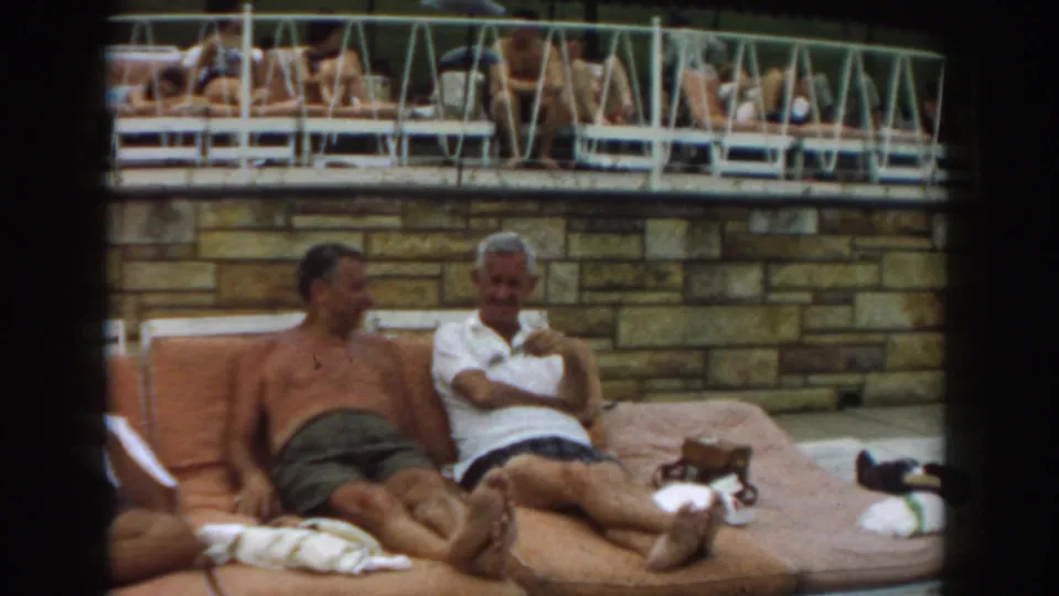 1961 Los Angeles frame of two men seated on orange poolside loungers, with a raised deck and other guests behind them.