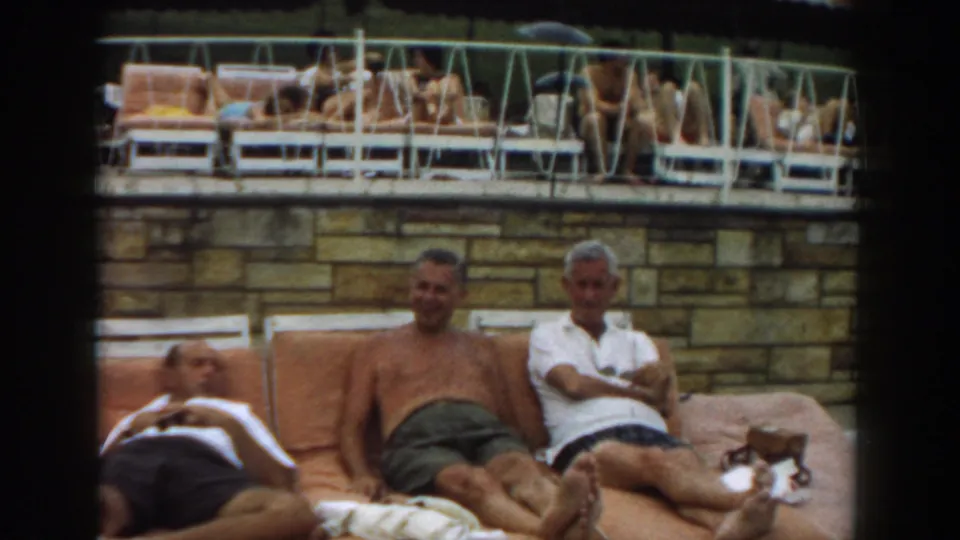 1961 Los Angeles home-movie frame with two seated men and a third reclined figure at the edge of a poolside lounge area.