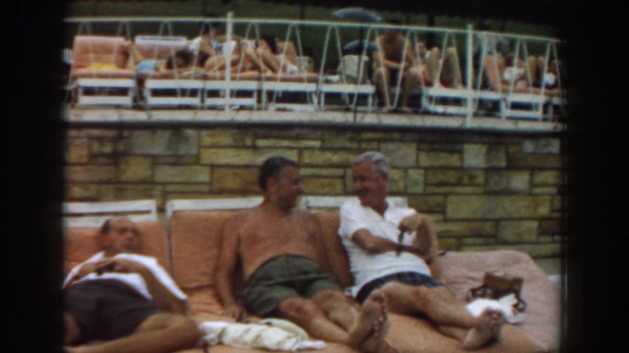 Three adult men reclining on orange poolside cushions with sunbathers behind a stone wall.