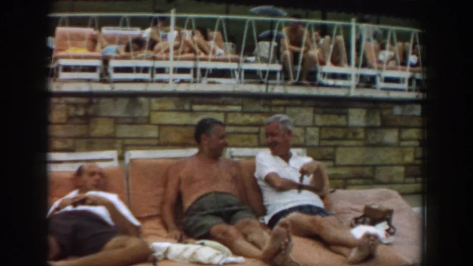 1961 Los Angeles home-movie frame of adult men relaxing on orange poolside cushions while guests lounge on an upper deck.