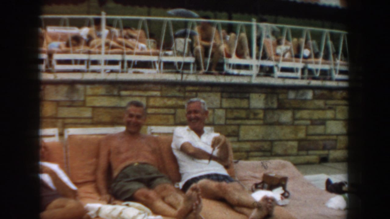 Two adult men seated on orange cushions at a poolside resort, with lounge chairs in the background.