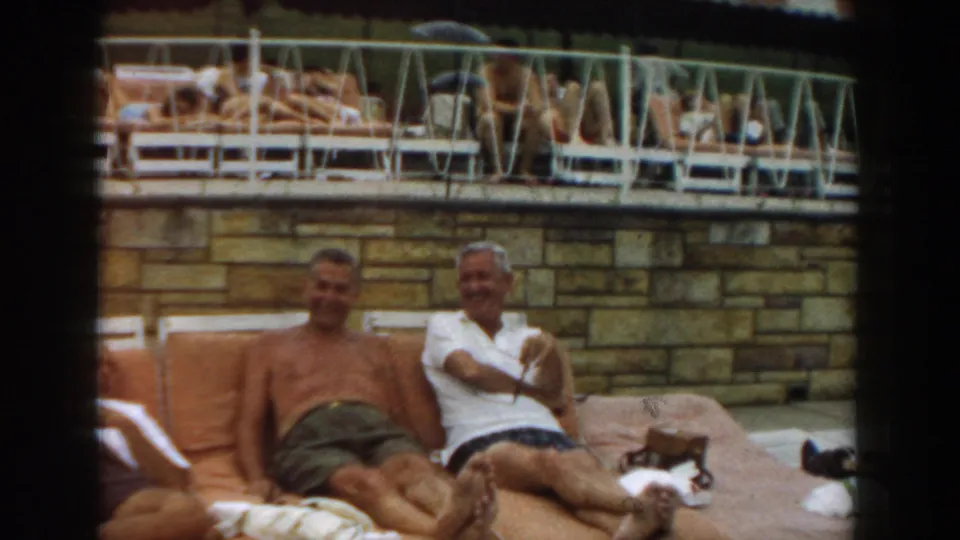 Two adult men smile and sit on orange poolside cushions in Los Angeles, with sunbathers visible on an upper deck.
