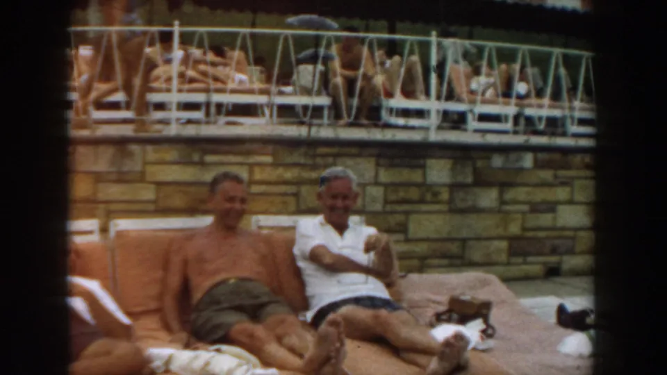 1961 Los Angeles poolside frame showing two men seated on orange cushions, with another reclining figure at the edge.