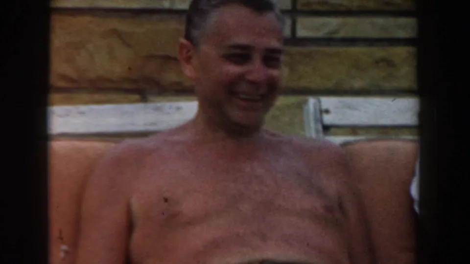 Close 1961 Los Angeles home-movie view of a smiling shirtless man seated on poolside cushions near a stone wall.