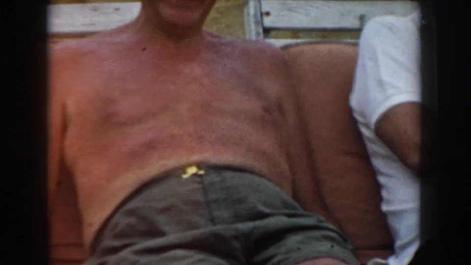 1961 Los Angeles home-movie frame of a shirtless older man sunbathing on cushioned outdoor seating beside another person.