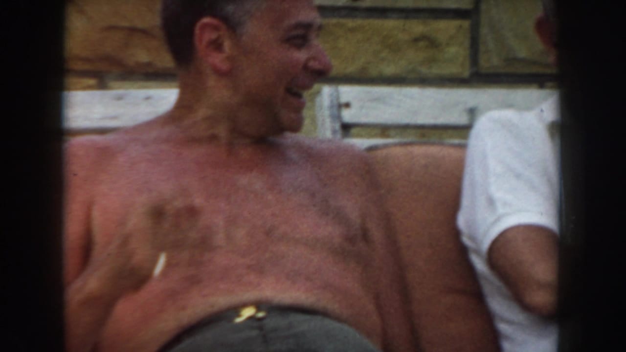 1961 Los Angeles frame showing a shirtless older man laughing or speaking while seated in bright sunlight.