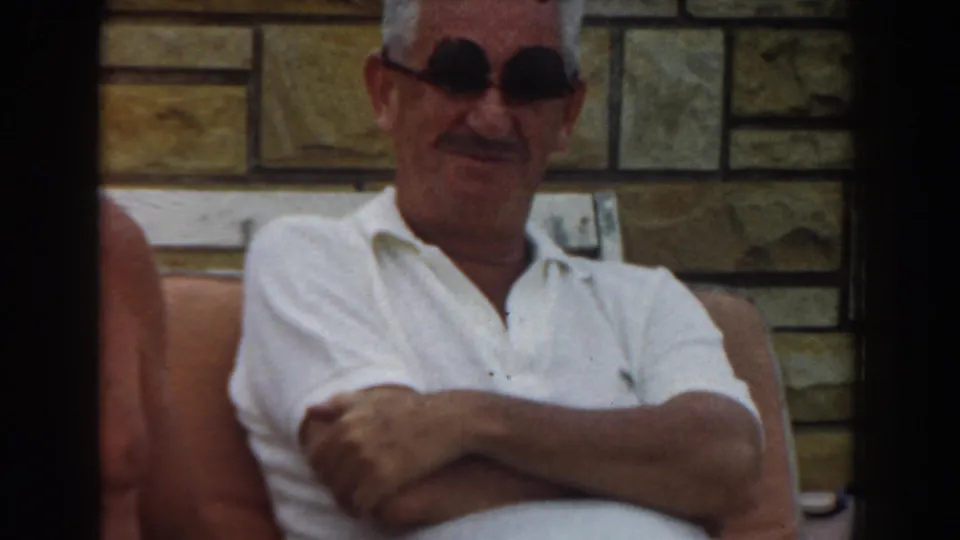 1961 Los Angeles home-movie frame of an older man in a white polo and dark sunglasses seated outdoors by a stone wall.