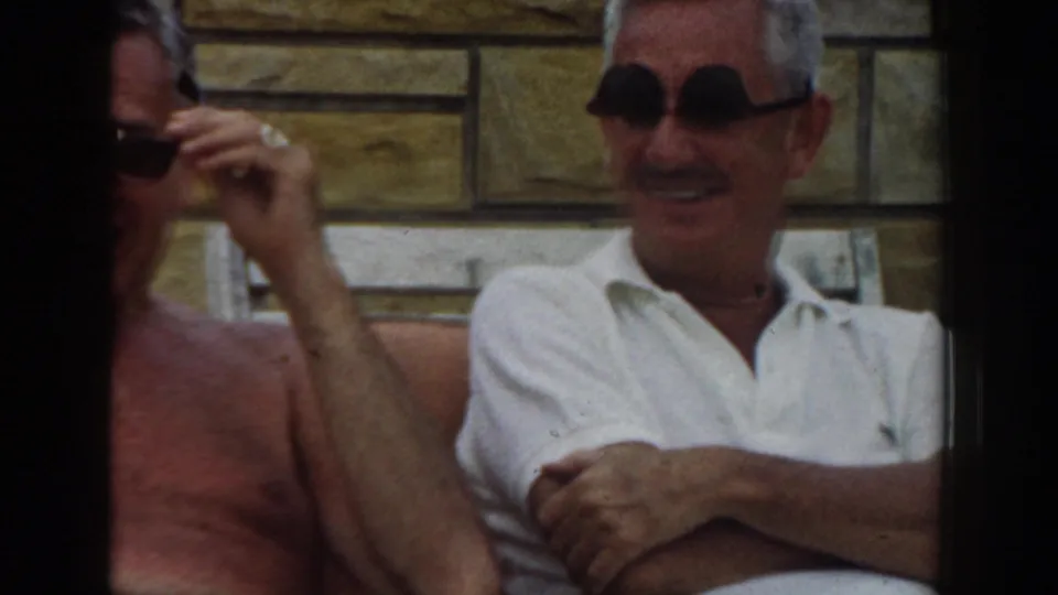 1961 Los Angeles frame of two older men seated together outdoors, one shirtless and one in dark sunglasses and a white polo.