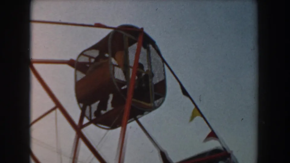 1961 Los Angeles 8mm frame showing a red Ferris wheel gondola with passengers silhouetted behind mesh against a pale sky.