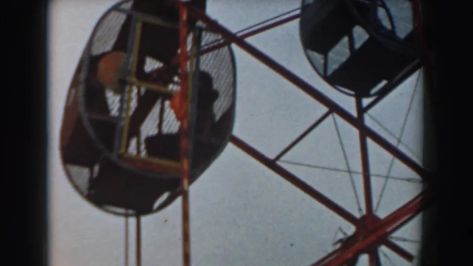 Close 8mm frame of a red Ferris wheel gondola with seated riders behind mesh and another car partly visible nearby.