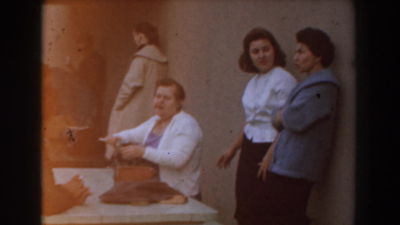 Adults near a table, with one woman seated and others standing nearby.