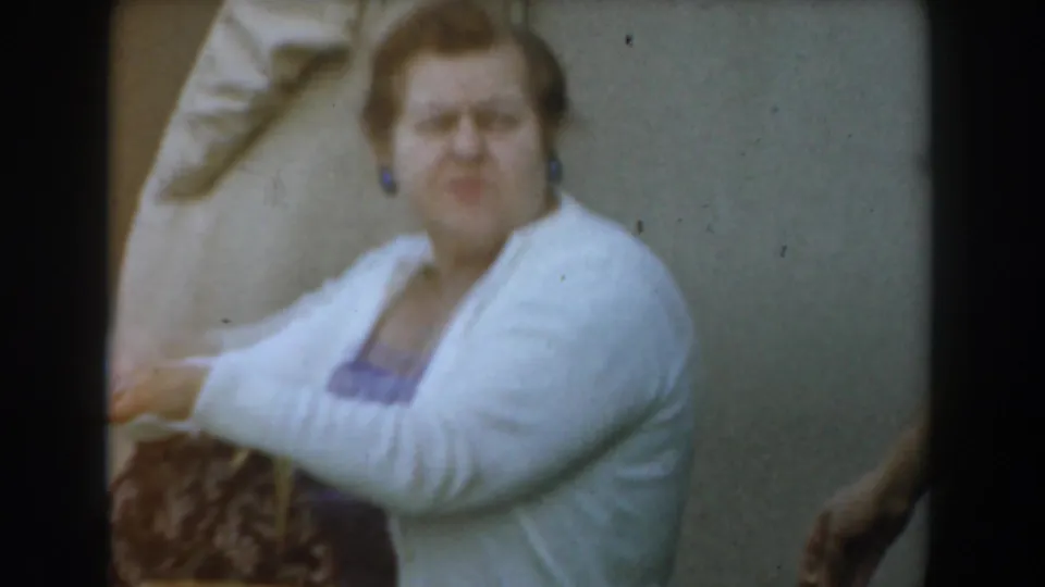 Close 1961 Los Angeles frame of an adult woman in a white cardigan seated beside a table, looking toward the camera area.