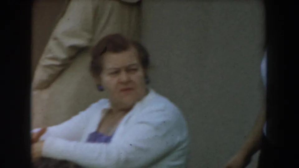 1961 Los Angeles home-movie frame with a seated adult woman in a white cardigan looking to the side while others stand at the edge of view.