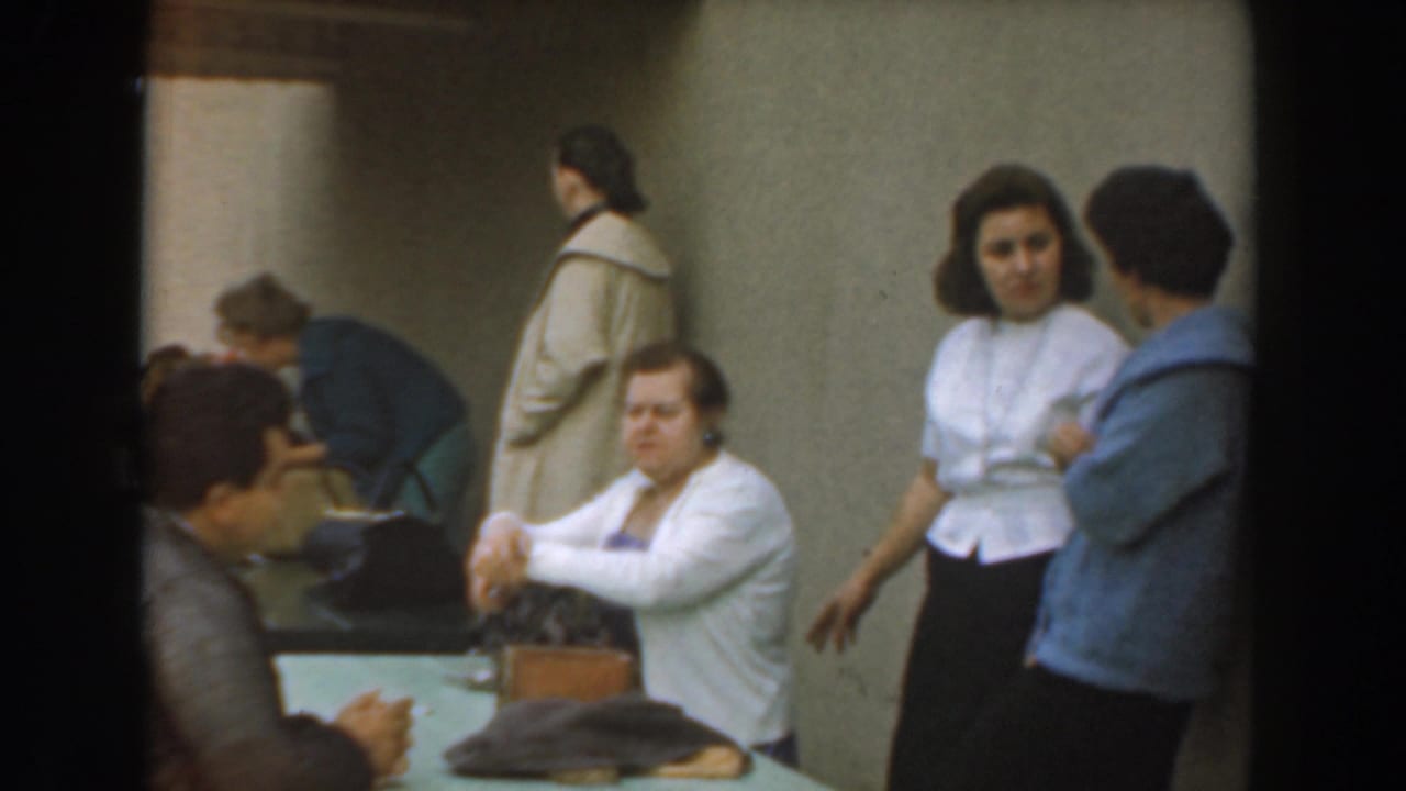 Adults sit and stand around a table in a plain room, with two women talking at right.