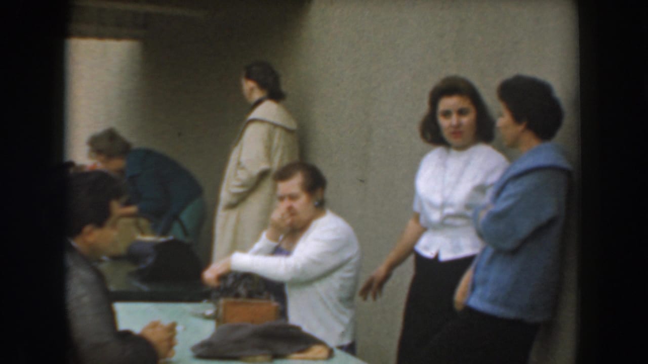 Candid 1961 Los Angeles indoor scene with a seated man, adults at a table, and two standing women conversing.