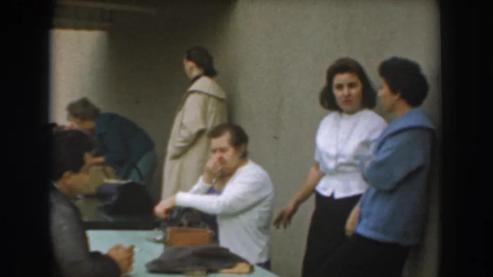 Candid 1961 Los Angeles indoor scene with a seated man, adults at a table, and two standing women conversing.
